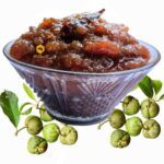 Glass bowl filled with sweet Chalta Chutney topped with red chili alongside fresh raw elephant apples on white background